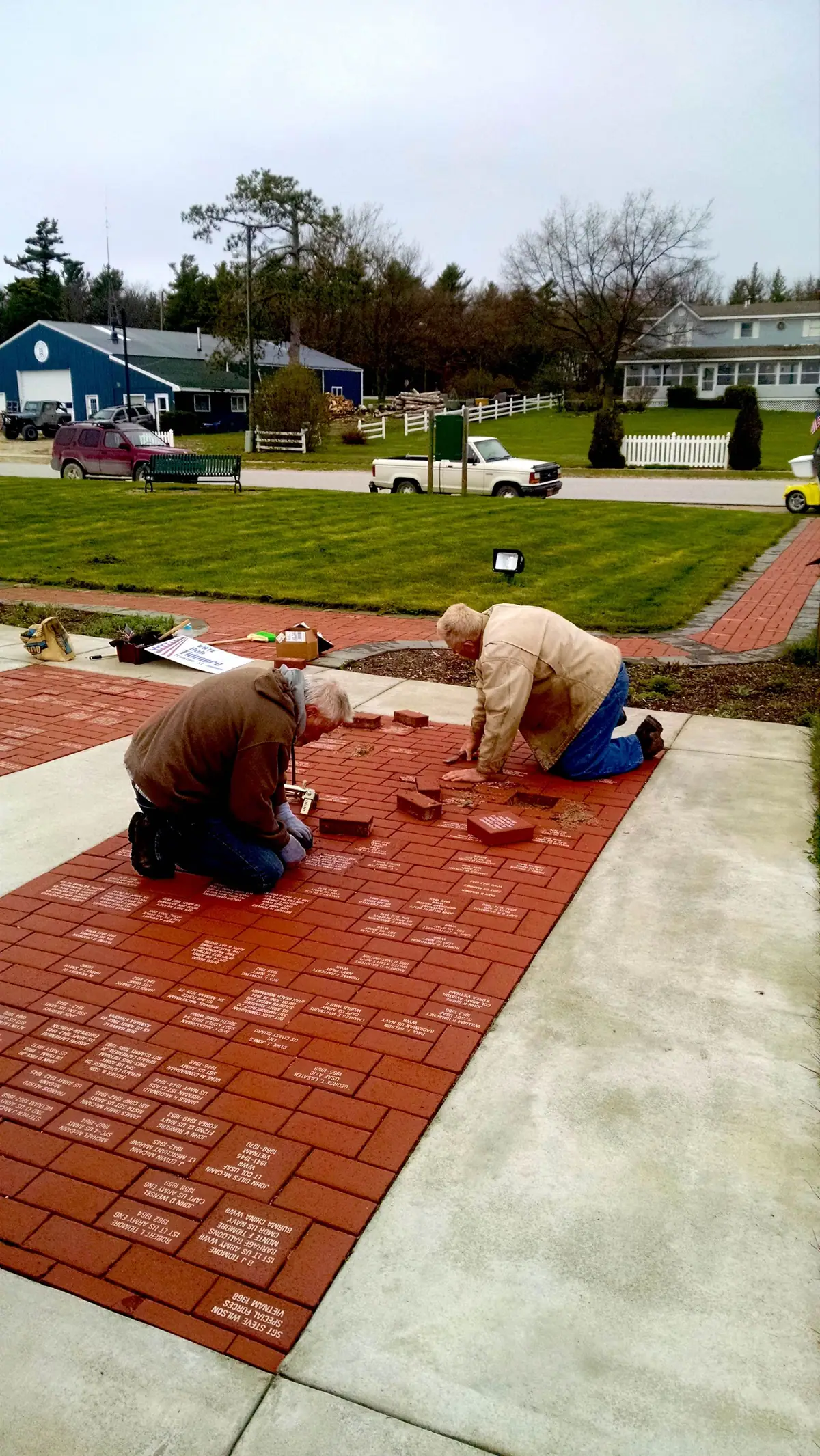 Two people lay red engraved bricks in a memorial walkway beside a lawn.
