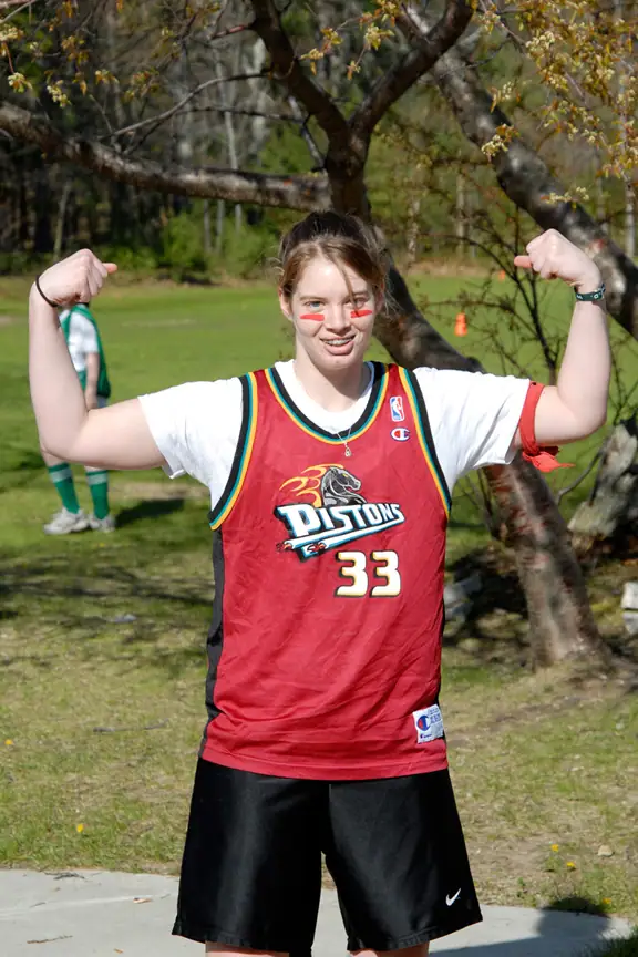 A smiling young woman flexes both arms while wearing a red basketball jersey and red face paint.