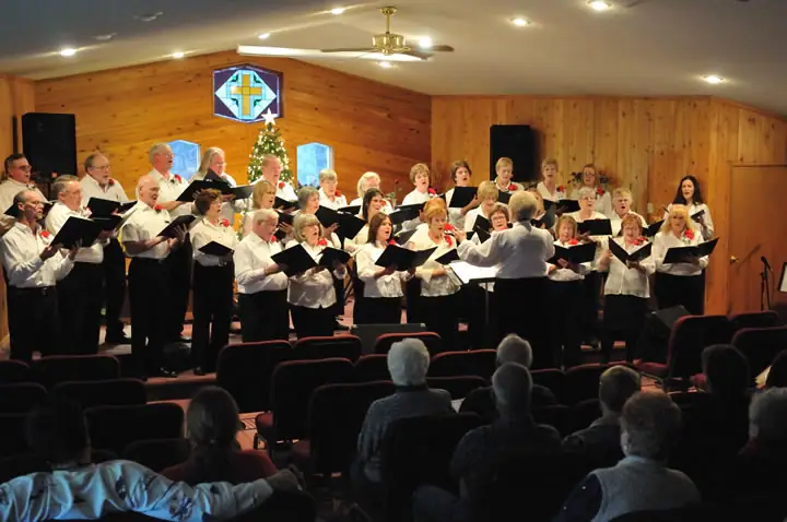 A choir performs before a seated audience in a wood-paneled hall.
