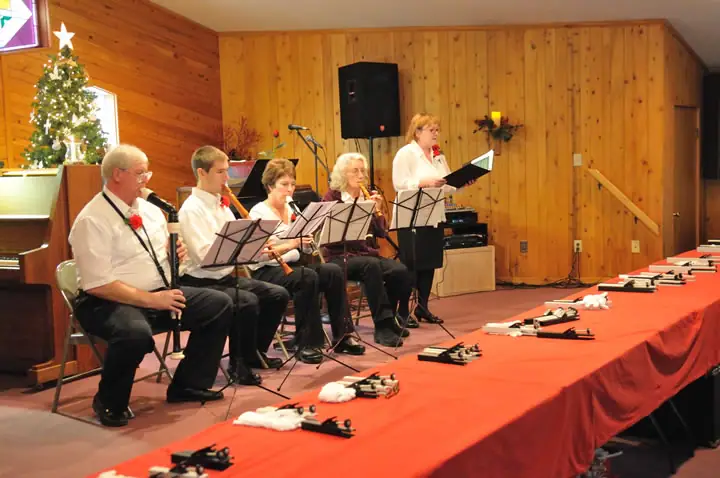 Five musicians perform indoors beside a decorated Christmas tree, with woodwind instruments displayed on a long red-covered table.