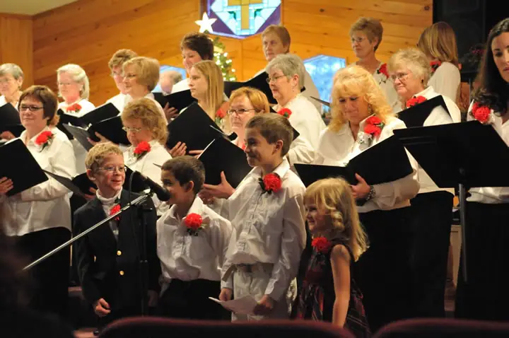 A choir and several children perform inside a wood-paneled church beneath a stained-glass window.