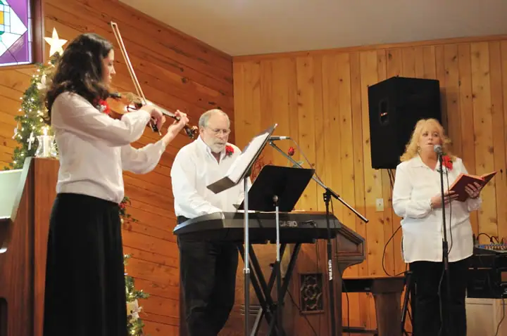 Three musicians perform in a wood-paneled room with a violin, electronic keyboard, microphone, and Christmas tree.