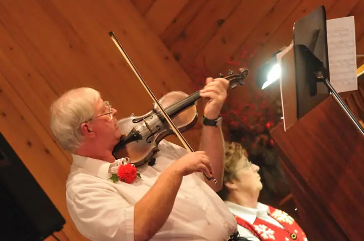 A violinist performs beside a music stand while another person looks upward in a wood-paneled room.