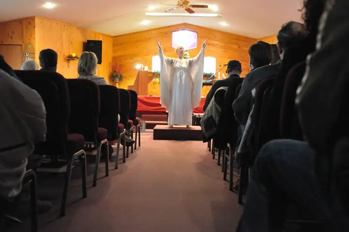 A robed officiant raises both arms before seated attendees in a wood-paneled church.