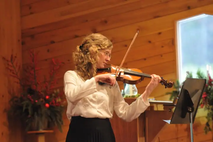 A violinist performs beside a music stand in a warmly lit wood-paneled room.