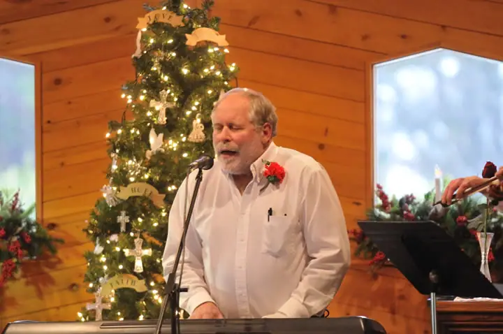 A singer performs beside a decorated Christmas tree while another musician plays at the right.