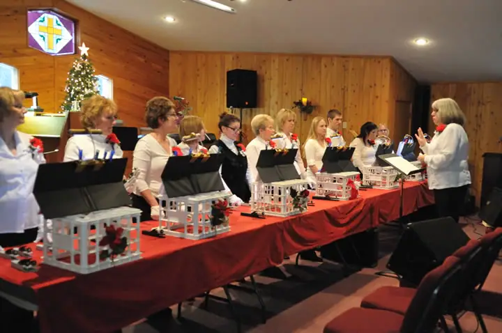 A group of musicians performs behind a decorated table while a conductor directs them in a wood-paneled room.