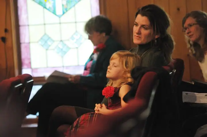 A child and three adults sit in a wood-paneled interior beside a stained-glass window.
