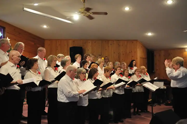 A choir sings from open music folders while a conductor leads them in a wood-paneled room.