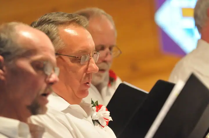 Several men in white shirts sing from black music folders, with flowers pinned to some shirts.