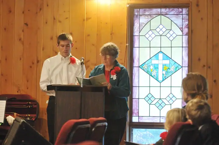Two people read from a book at a podium before a seated audience in a wood-paneled interior.