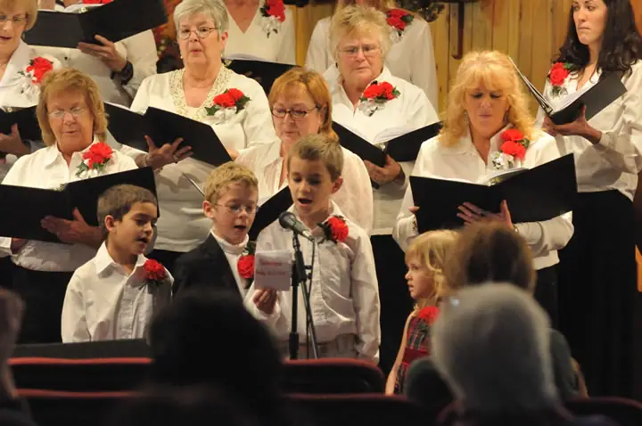 A choir in white performs with children at microphones before an audience.