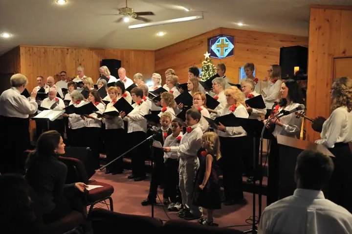 A choir performs with a conductor in a wood-paneled room decorated with a cross and Christmas tree.