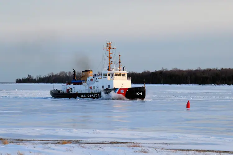 A Coast Guard cutter breaks through ice near a wooded shoreline, with a red buoy in the foreground.