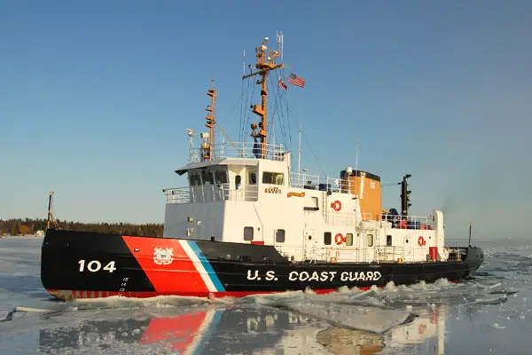 A Coast Guard vessel marked 104 moves through broken ice near a wooded shoreline.