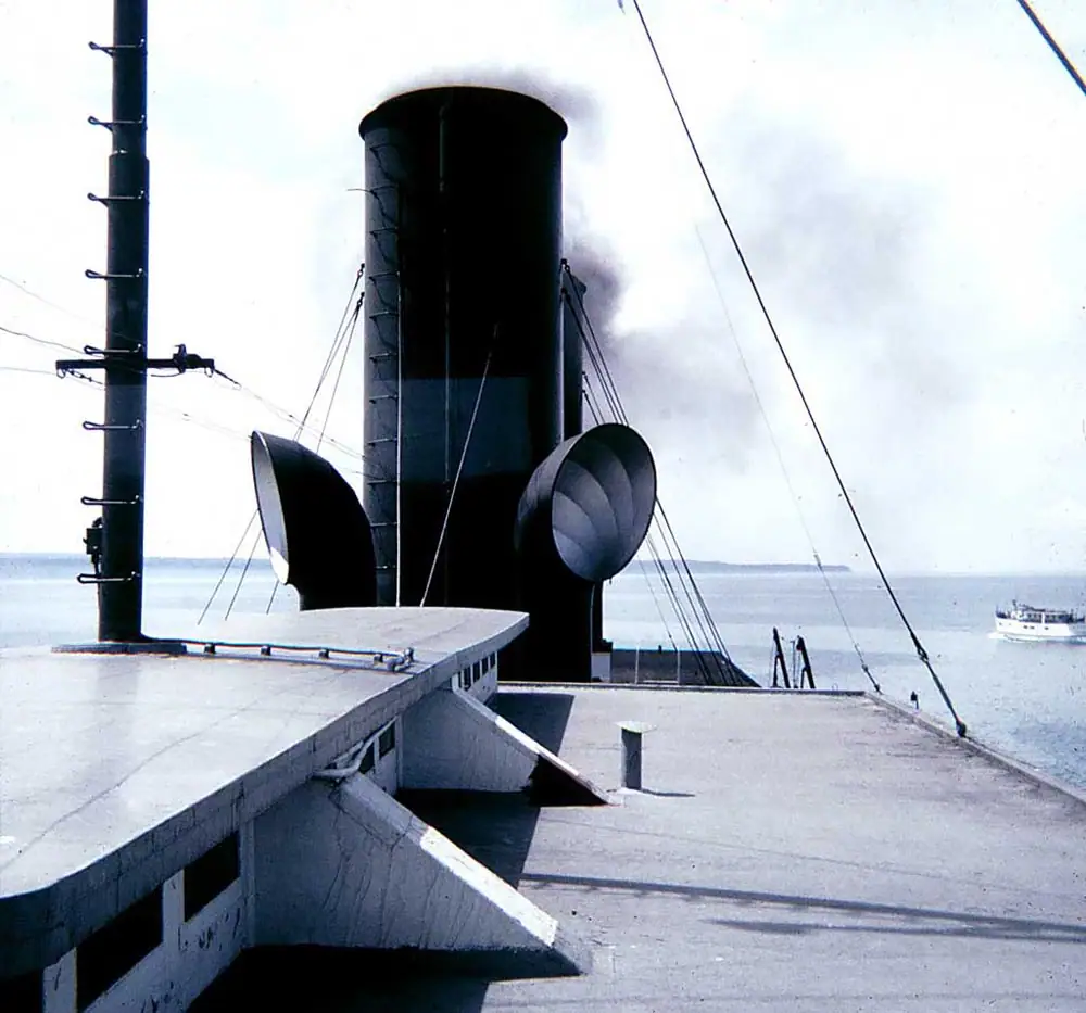 Ship deck with a large black smokestack, mast, rigging, and a small boat on the water beyond.