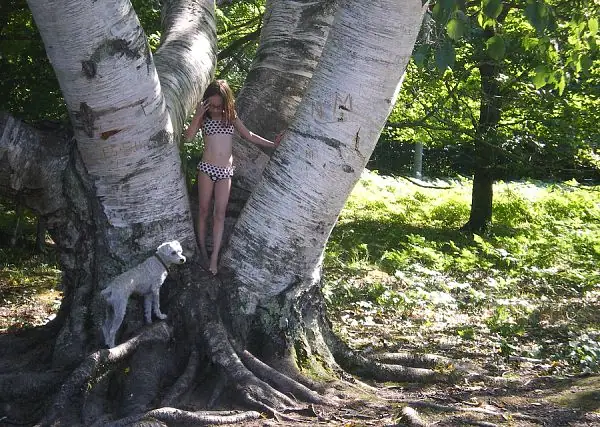 A child in a polka-dot swimsuit stands beside a white-barked birch tree with a small white dog.