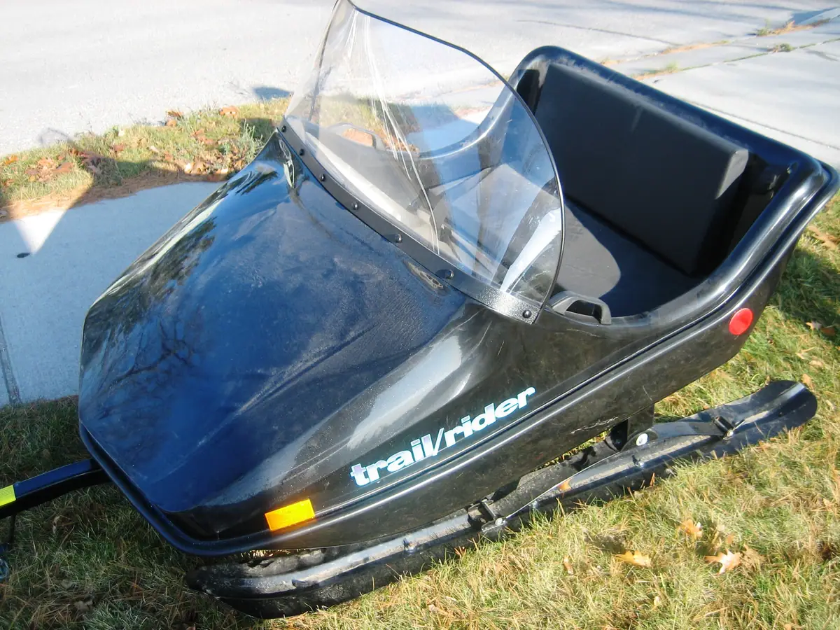 Black snowmobile with a clear windshield parked on grass beside a concrete sidewalk.