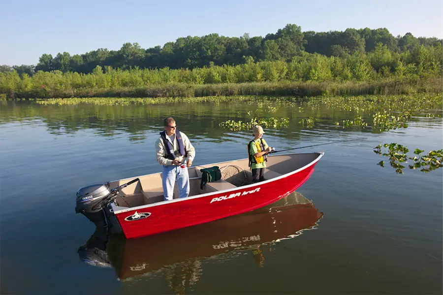 An adult and child fish from a red motorboat on calm water beside a tree-lined shore.