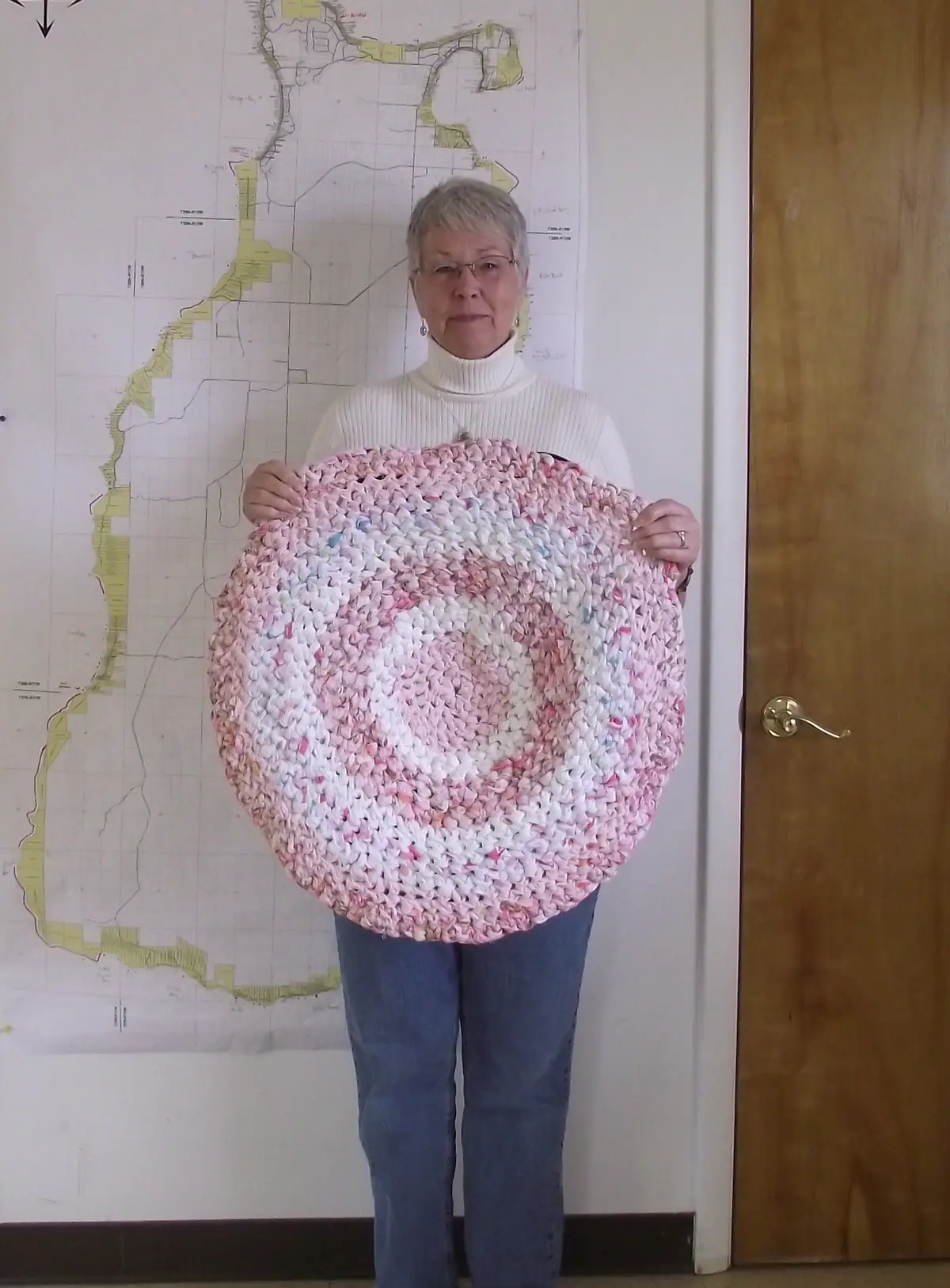 A woman holds a large round crocheted rug in front of a wall map.