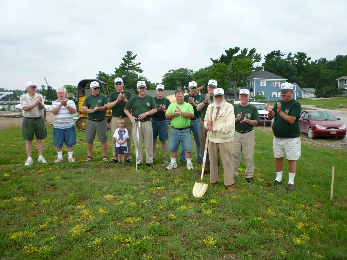 A group applauds beside a ceremonial shovel on a grassy lot with houses, cars, and a waterfront behind them.