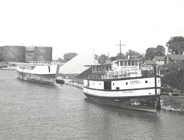 Black-and-white photograph of two boats moored beside a shoreline, with silos, houses, and a mail boat marked NORTH SHORE.
