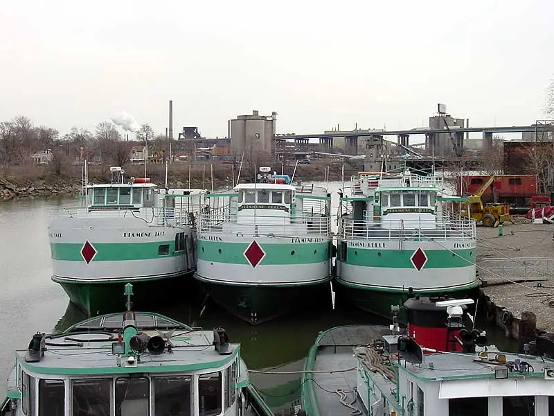 Three green-and-white vessels are moored side by side at an industrial waterfront, with two more boats in the foreground.