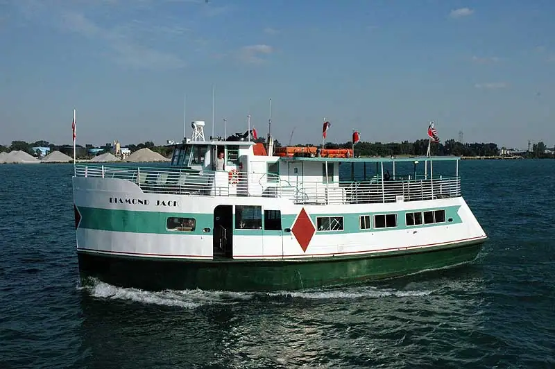 A green-and-white passenger ferry travels past a shoreline with pale piles of material.