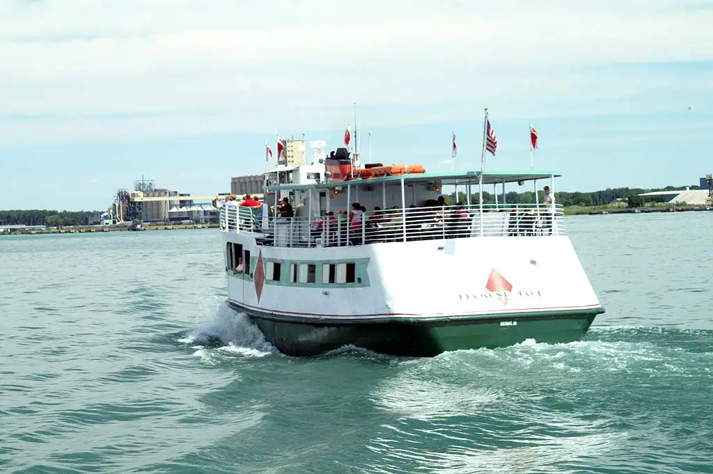 A white-and-green passenger ferry carries people past an industrial waterfront, with flags flying above its decks.