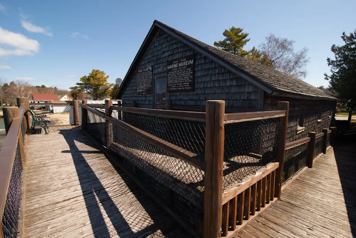 Shingled marine museum building on a fenced wooden pier, with benches and interpretive signs.