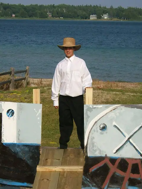 A young man in a straw hat stands on a wooden walkway between painted boat-like panels beside the water.