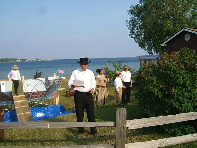 People in period-style clothing gather beside a decorated boat display near the water.