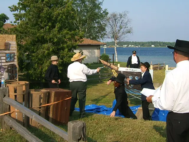People in period-style clothing perform outdoors beside a model boat and waterfront.