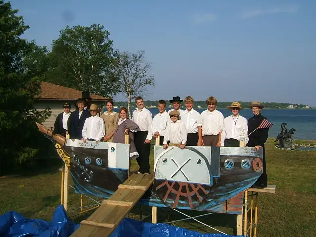 Thirteen people pose with a brightly painted wooden boat model beside the water.