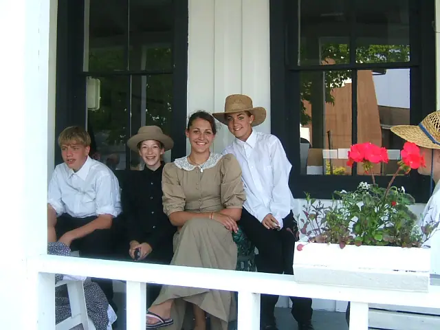 Four young people in period-style clothing sit on a porch beside flower boxes.