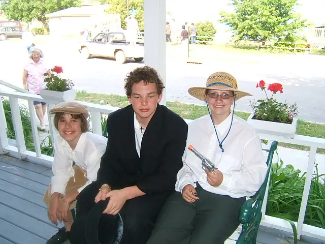 Three people pose on a porch beside flower boxes, with parked cars and houses visible behind them.