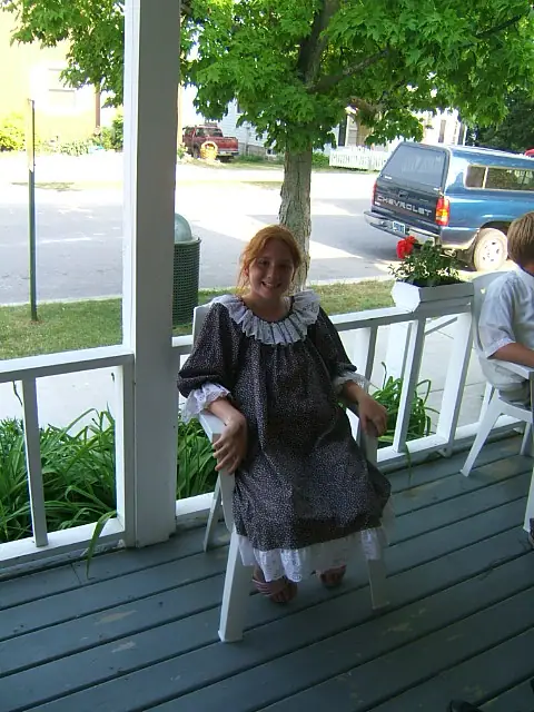 A girl in a dark historical-style dress sits on a porch beside a street and a blue pickup truck.