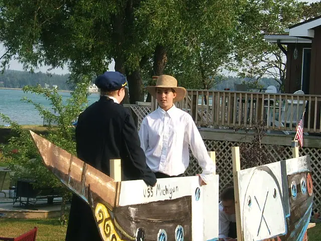 Two young people in sailor-style costumes stand behind a painted wooden boat-shaped structure beside the water.