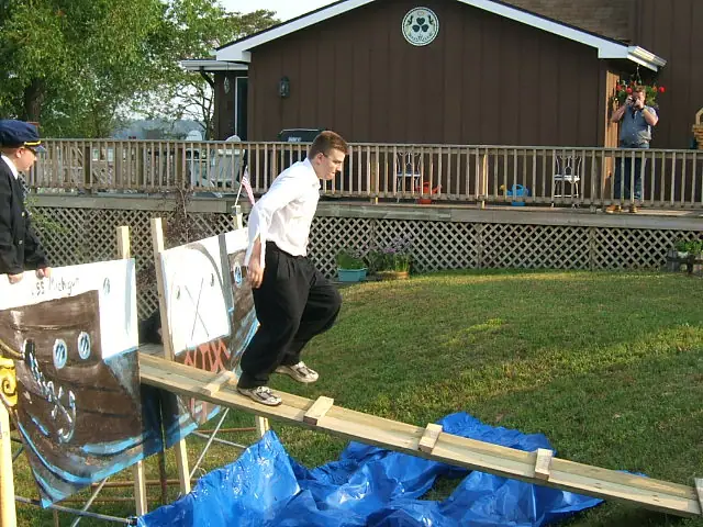 A person balances on a wooden plank over a blue tarp beside a porch while others watch and photograph the scene.