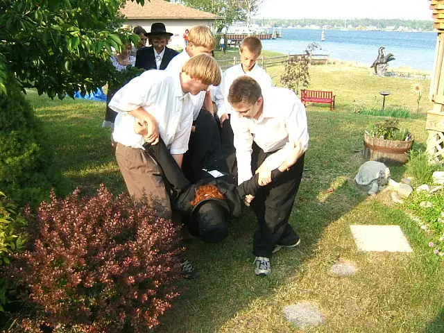 Several people carry a black-clad figure across a grassy waterfront garden near a dock and small boats.