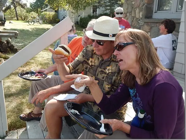 Adults eat outdoors on the steps of a house, with plates, sandwiches, and bicycles visible nearby.