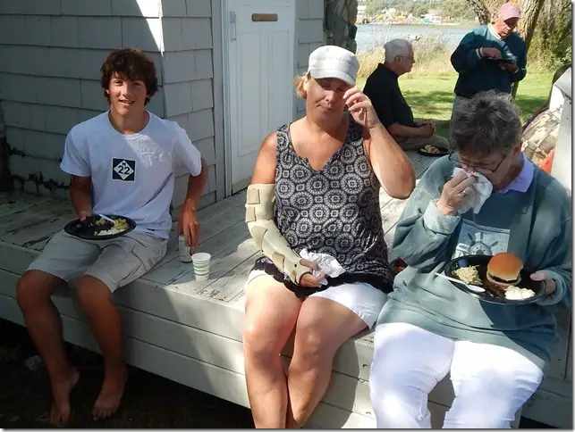 Three people eat outdoors on a bench while two others sit behind them near the water.
