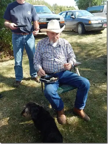 Two men gather outdoors as one eats from a plate beside a dog and parked cars.