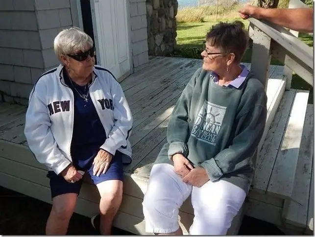 Two women sit on a wooden deck, with printed lettering visible on their clothing.