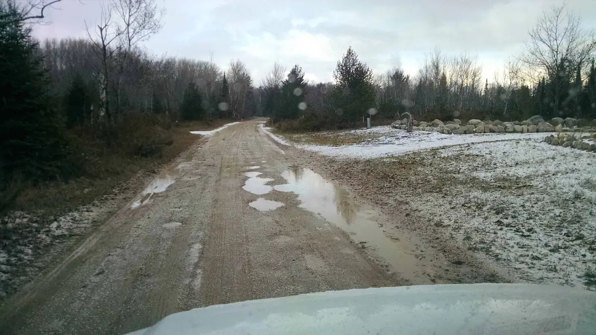 A muddy gravel road with puddles leads through a snowy forest past a pile of large rocks.