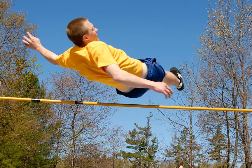 Athlete in a yellow shirt clears a yellow high-jump bar against a blue sky and trees.