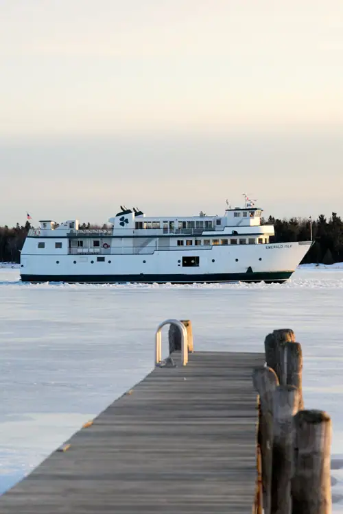A white ferry passes behind a wooden pier across a frozen waterway.