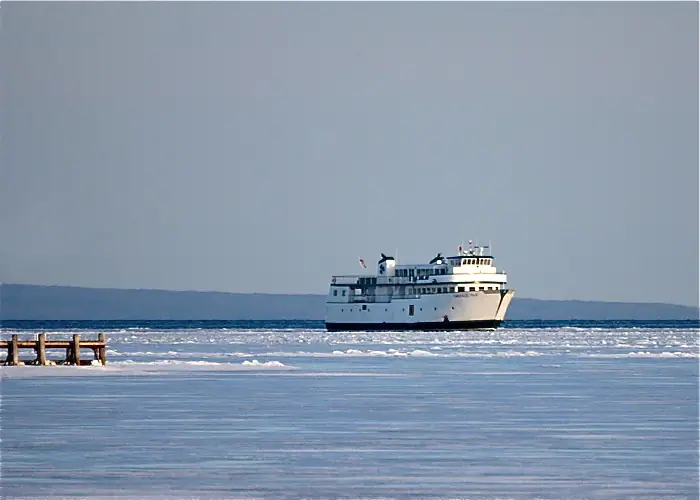 A white ferry moves through broken ice beyond a wooden pier under a gray sky.