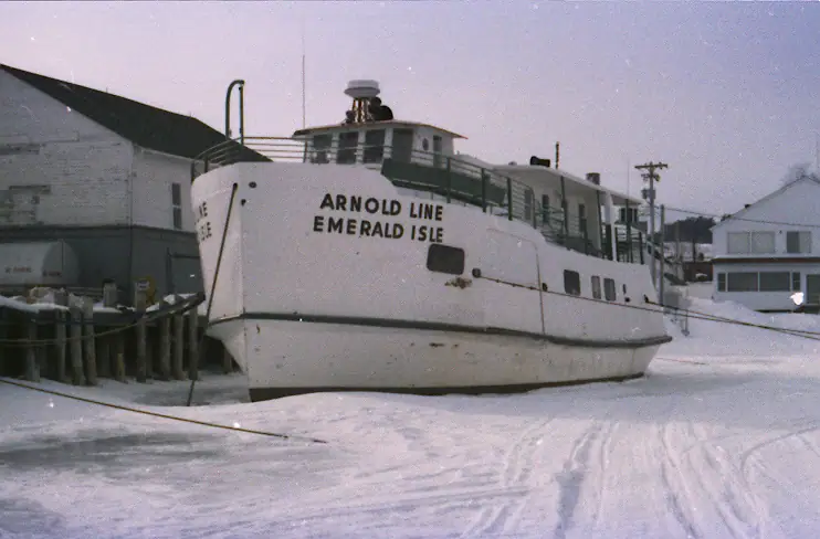 A white ferry marked “ARNOLD LINE EMERALD ISLE” sits beside a snowy dock and waterfront buildings.