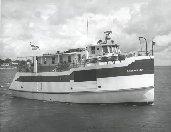 Black-and-white photograph of a ferry moving across the water with people on deck.